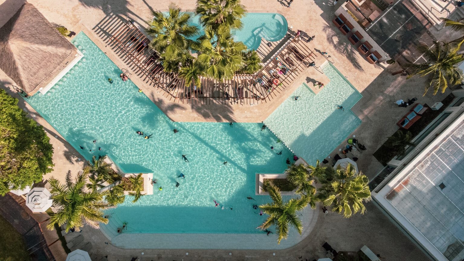 Aerial view of a luxurious outdoor pool area at The Husna Experience, featuring palm trees, lounge chairs, and a clear swimming pool in a vibrant resort setting.