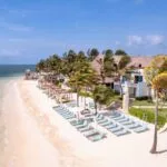 Aerial view of a tropical beach resort with white sand, turquoise waters, palm trees, and sun loungers under umbrellas, showcasing a luxurious seaside getaway.