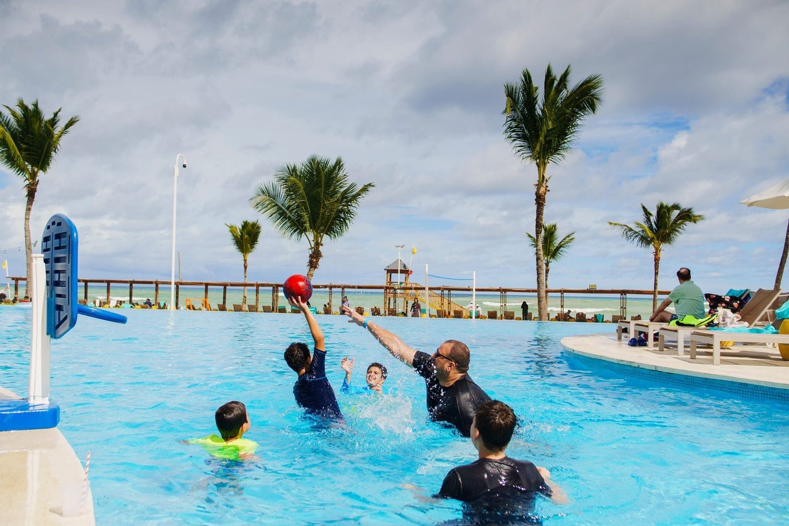 Family enjoying a lively game of water volleyball in a tropical resort pool with palm trees and ocean views in the background.
