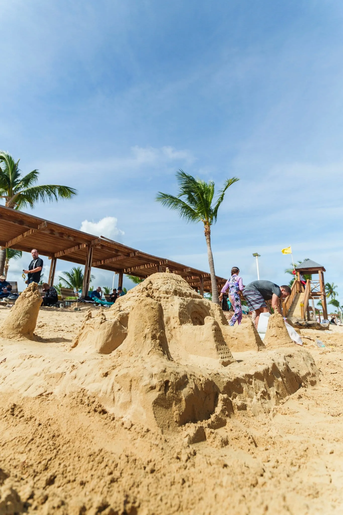 Vibrant beach scene showcasing a detailed sandcastle with palm trees and people enjoying the Husna beach experience under a clear blue sky.