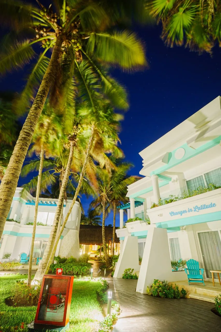 Relaxing tropical resort with illuminated white buildings and tall palm trees under a starry night sky.
