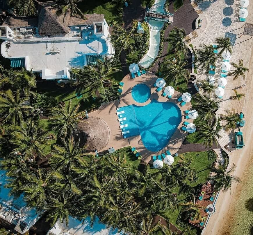 Aerial view of Husna Resort showcasing a large swimming pool surrounded by palm trees, sun loungers, and tropical landscaping, offering a luxurious and relaxing vacation experience.